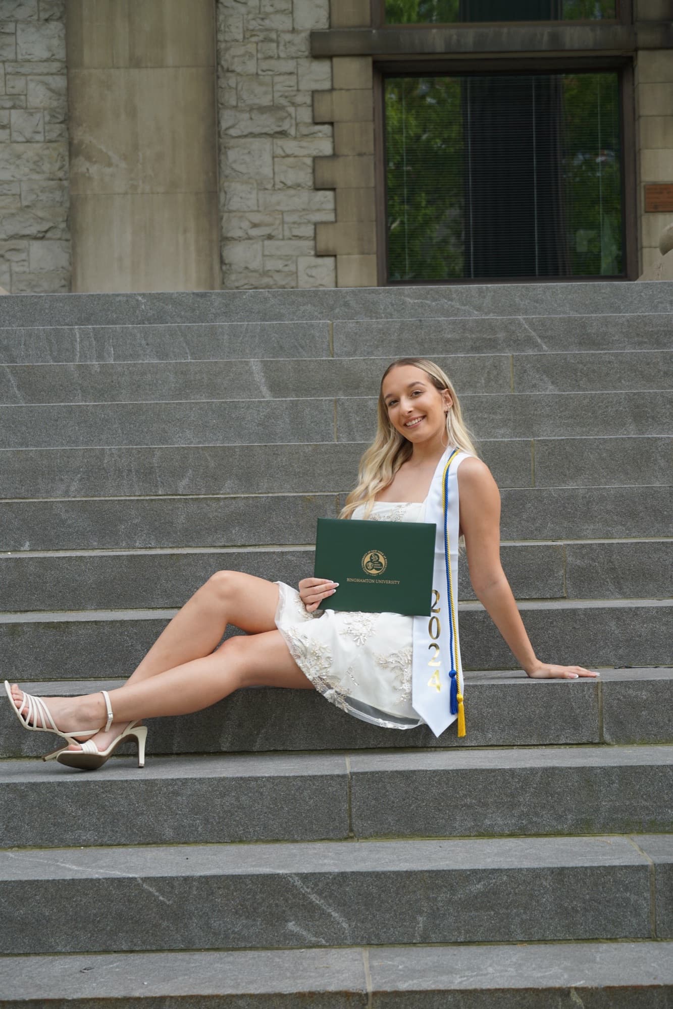 Graduate in a white lace dress and 2024 stole sitting on stone steps holding a Binghamton University diploma
