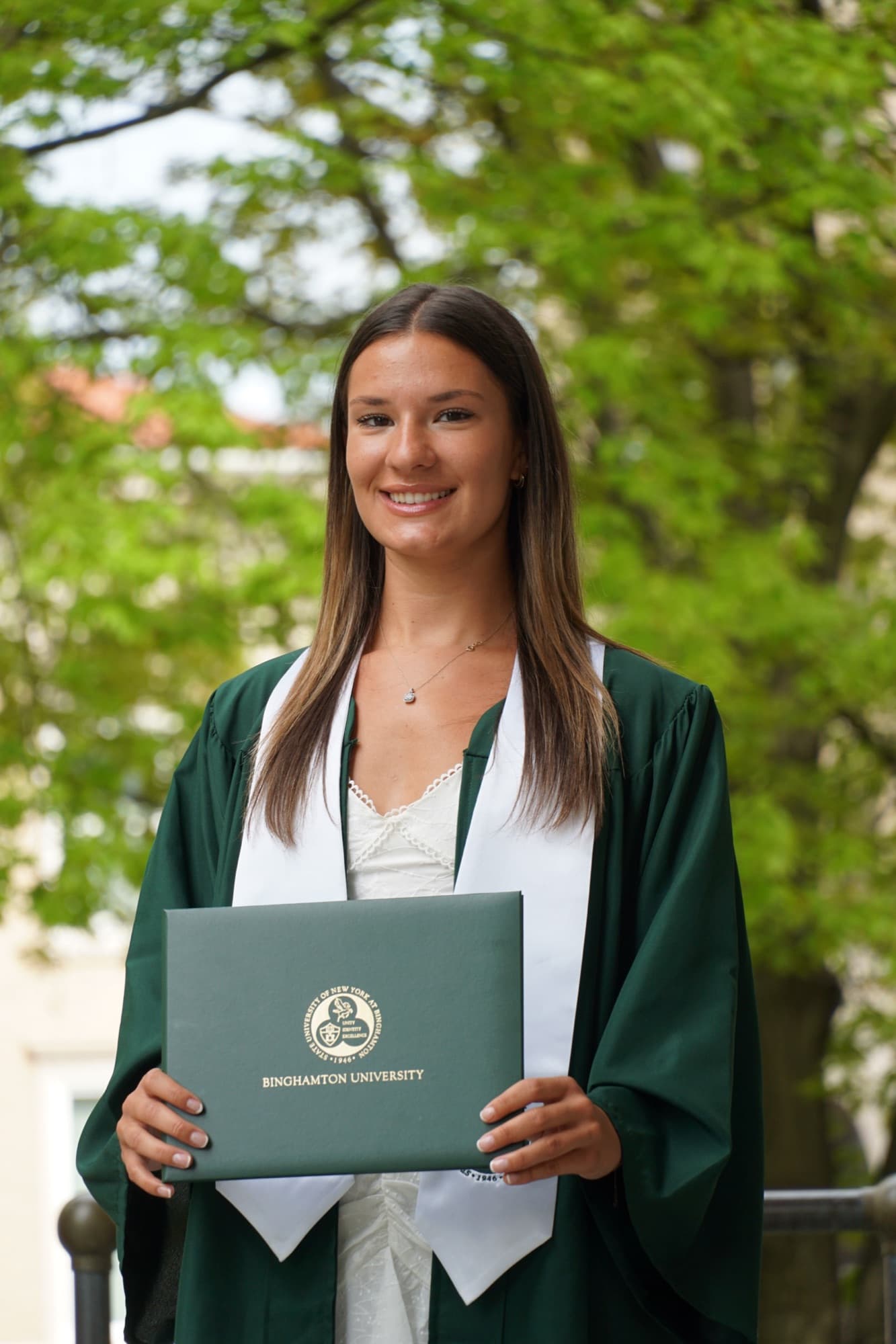 Smiling graduate in a green gown and white stole holding her Binghamton University diploma under spring trees