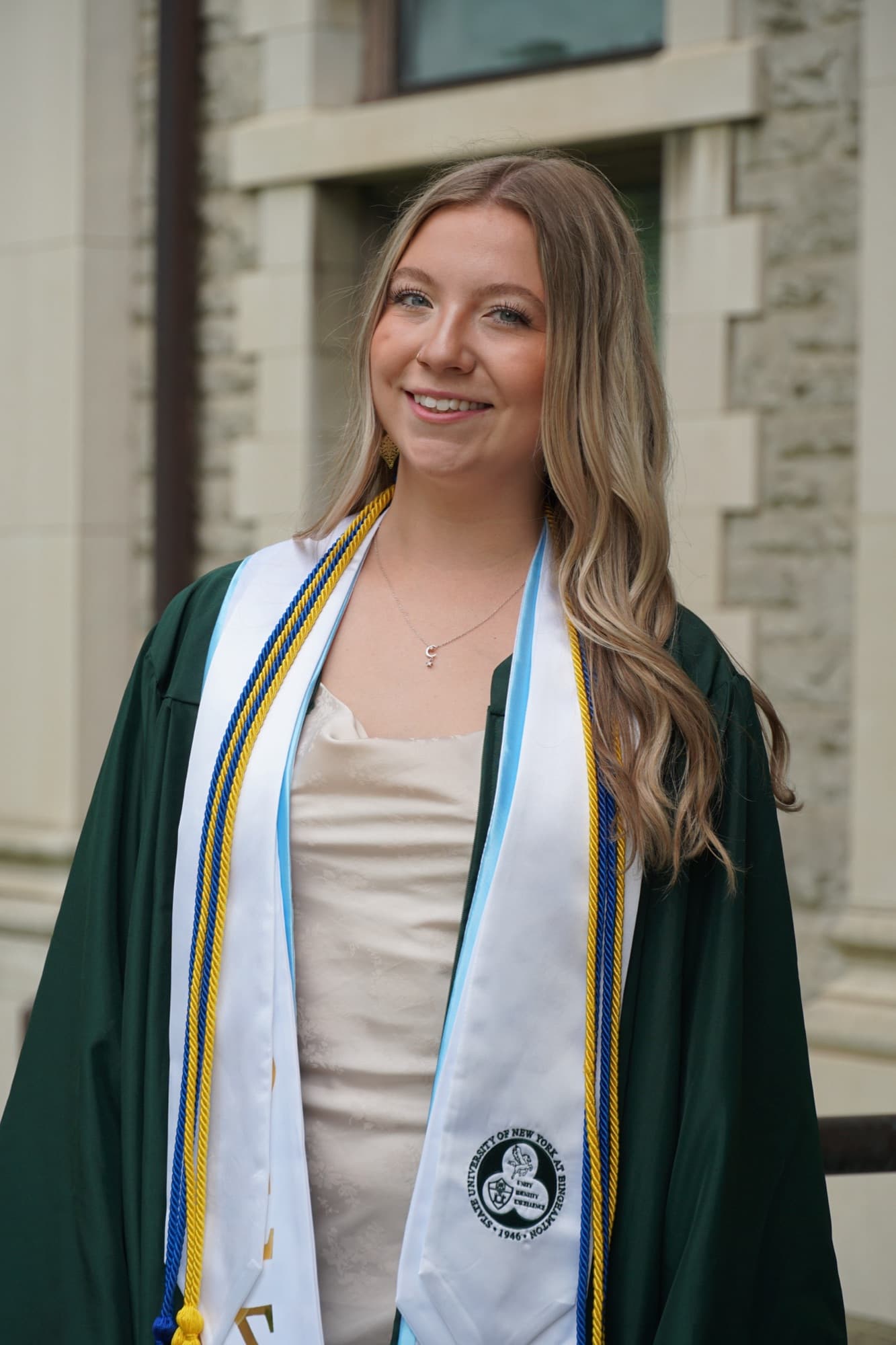 Graduate wearing honor cords and a SUNY Binghamton stole in front of a limestone building