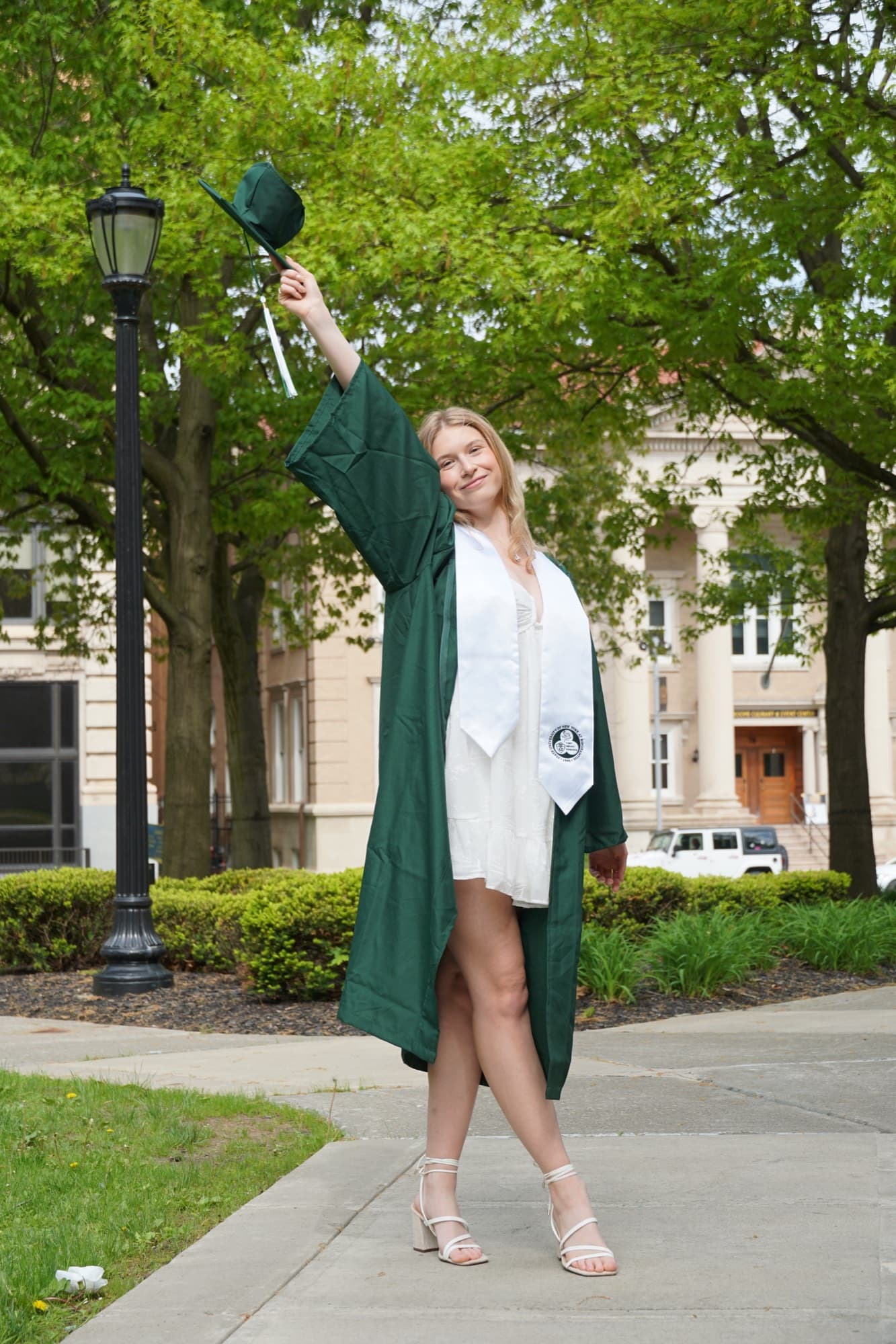 Graduate in an open green gown raising her cap on a tree-lined path in downtown Binghamton