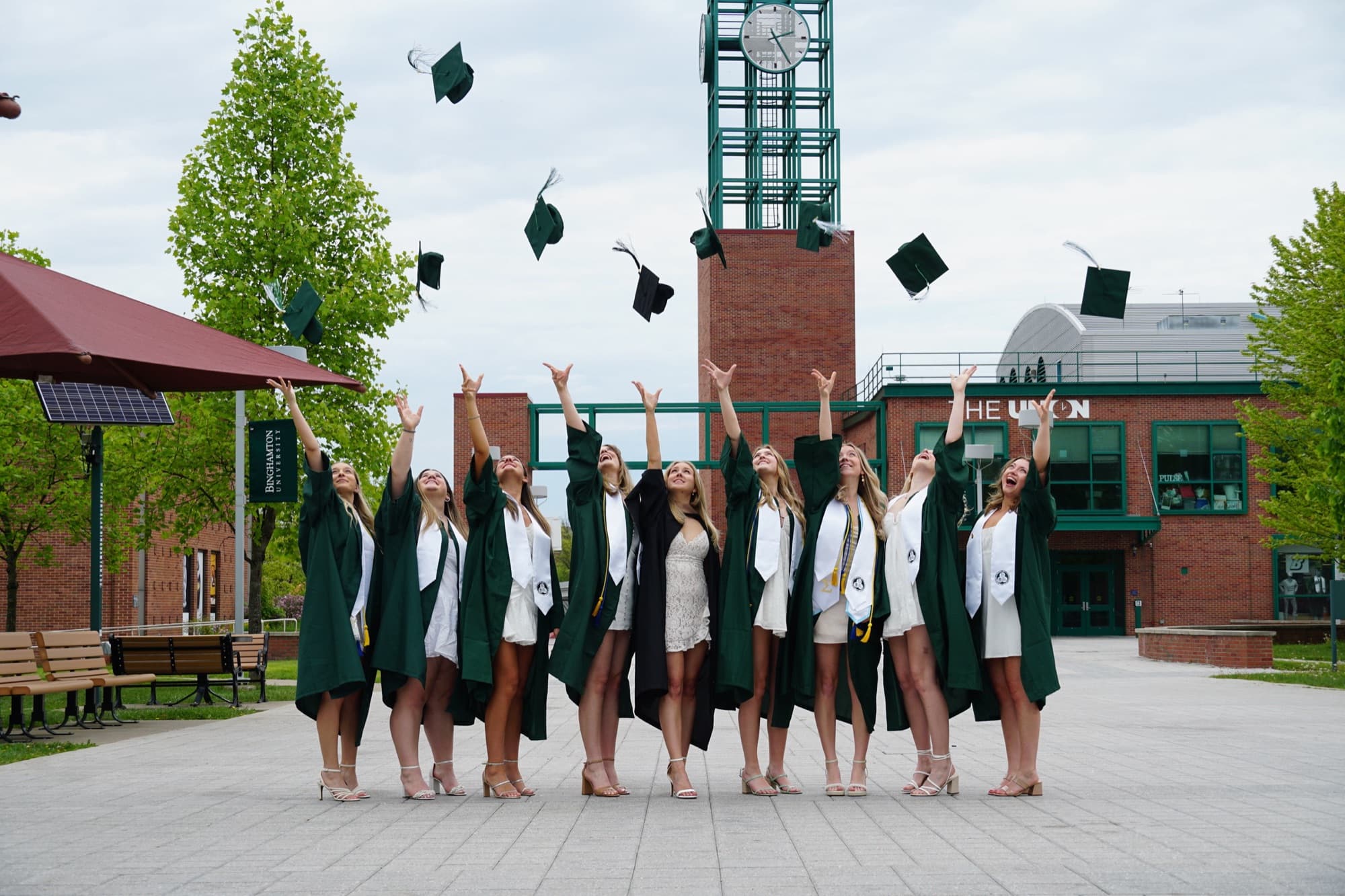 Nine Binghamton University graduates in green gowns tossing their caps in front of the Union clock tower