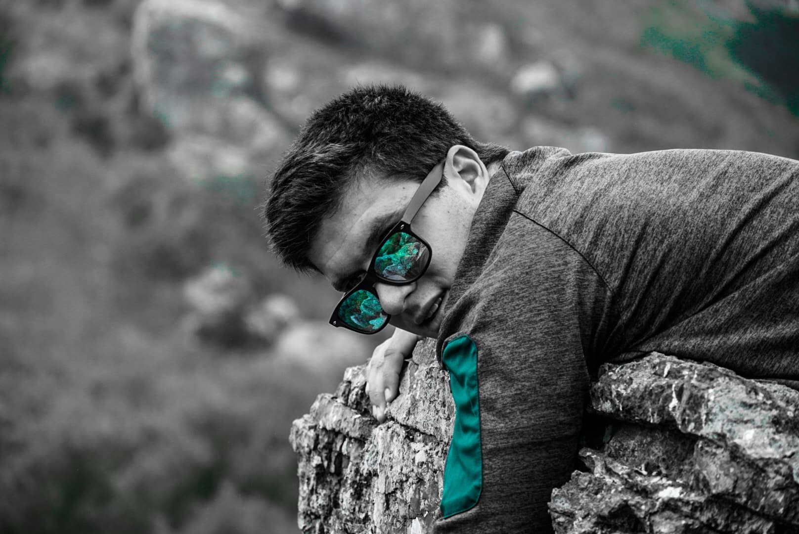 Selective-color portrait of a man in teal sunglasses leaning over a rock wall at a gorge overlook