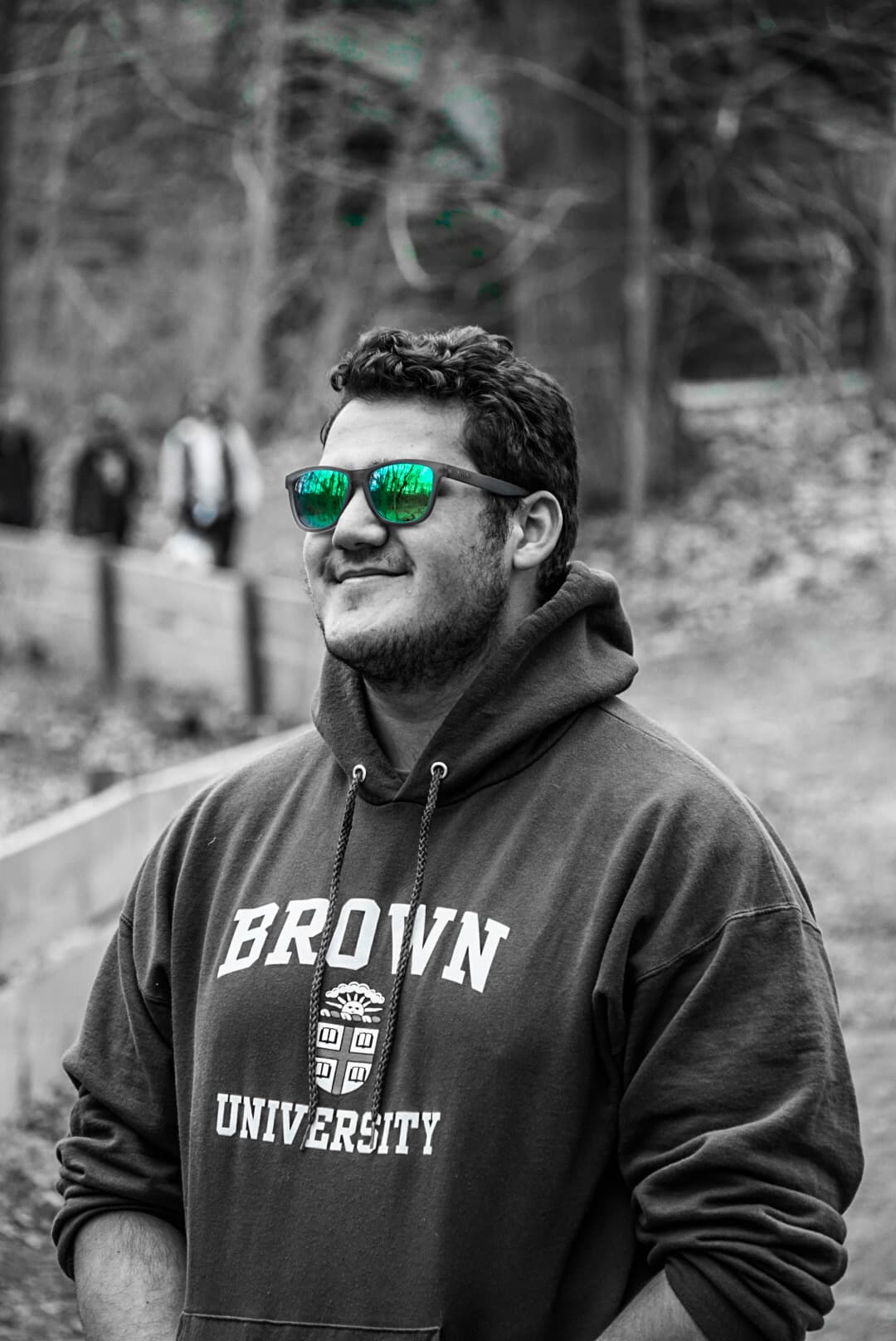 Selective-color portrait of Omar in green mirrored sunglasses and a Brown University hoodie on a wooded trail