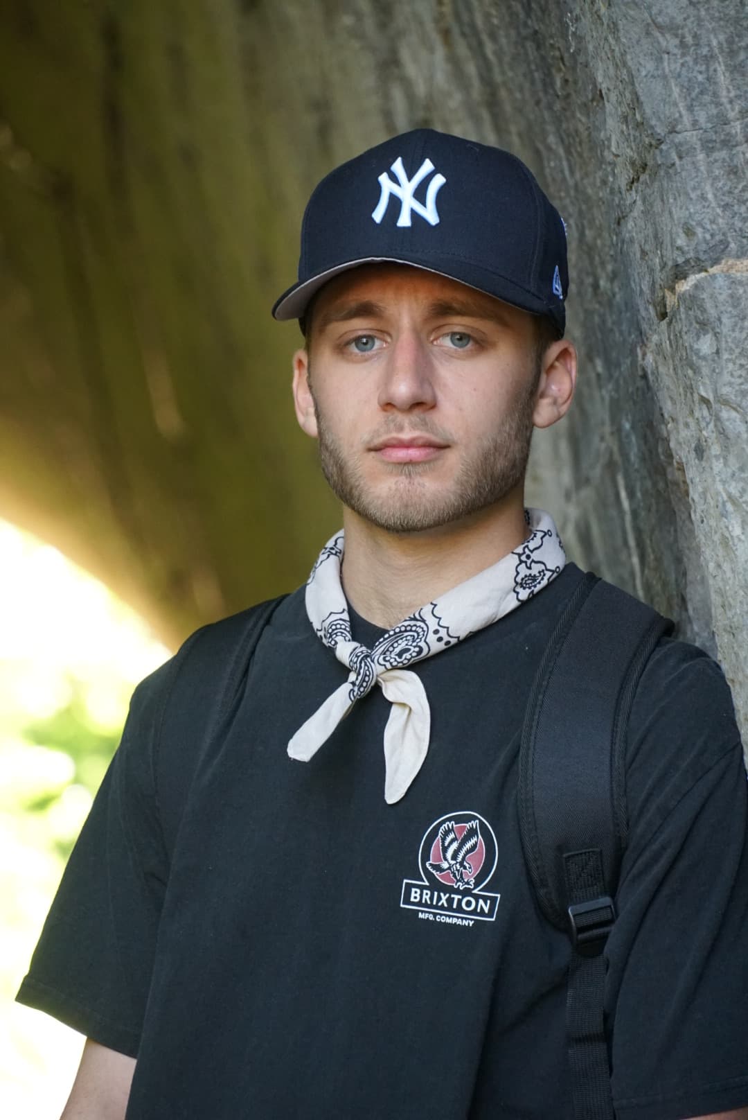 Solo portrait of Sam in a Yankees cap and bandana leaning against a stone tunnel wall
